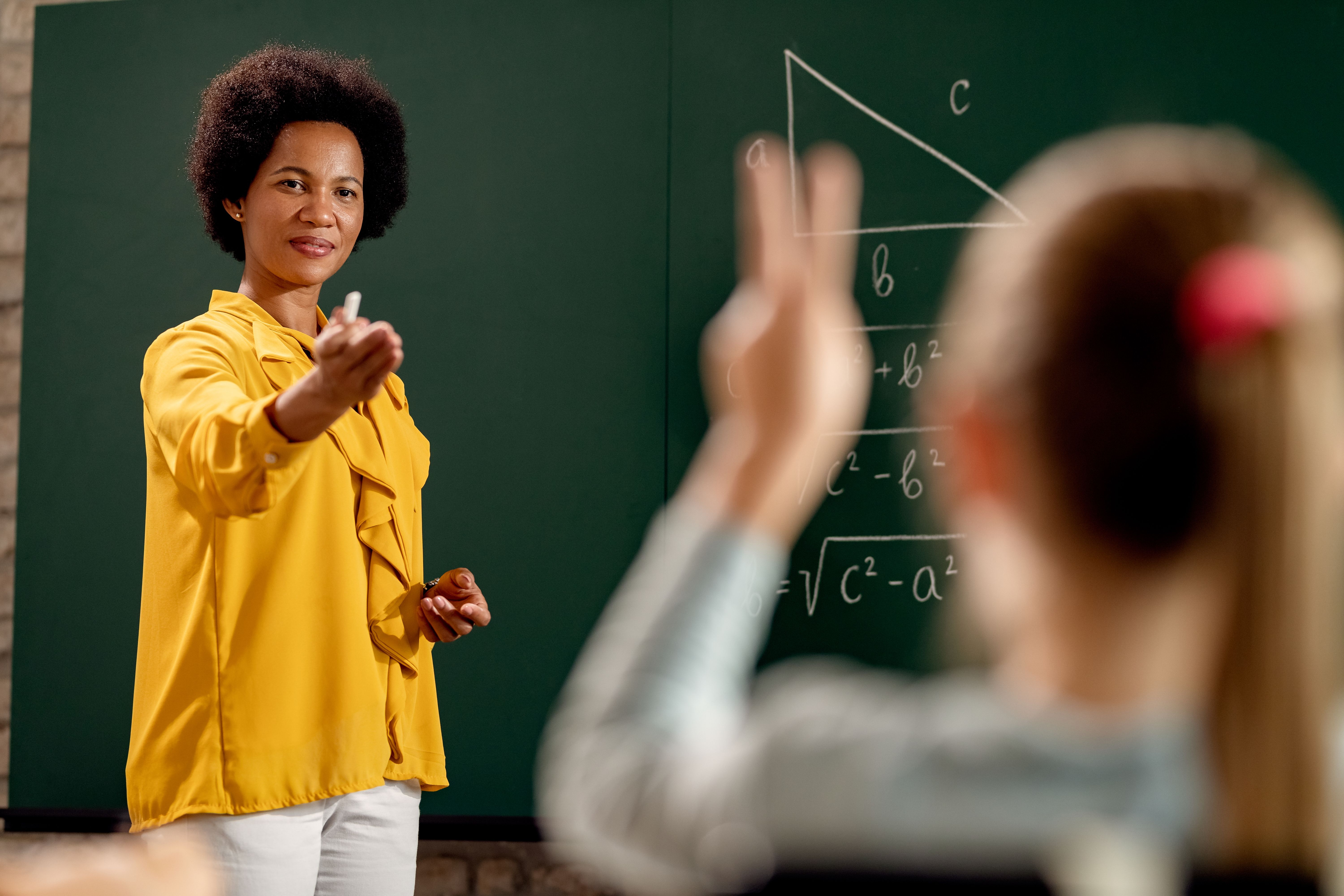 Professora substitua apontando para aluna com o braço levantado.